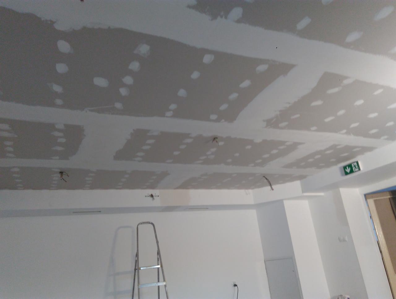 Montaż sufitu podwieszanego z płyt gipsowo-kartonowych, widoczne szpachlowane łączenia i wystające kable elektryczne, drabina aluminiowa oparta o ścianę, oświetlenie robocze i znak ewakuacyjny.