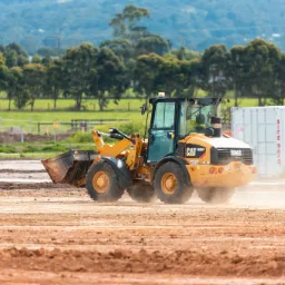 Żółta ładowarka CAT 906H podczas prac ziemnych na placu budowy, z widocznym kontenerem i drzewami w tle.