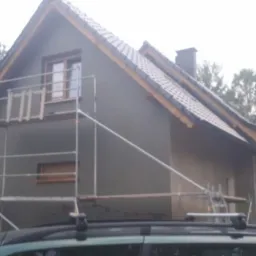 Dom jednorodzinny w trakcie ocieplania elewacji, widoczne rusztowania, szara elewacja i fragment samochodu na pierwszym planie.