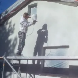 Pracownik w białej bluzie i spodniach moro, stojący na rusztowaniu, aplikuje białą farbę natryskowo na elewację domu. Okna zabezpieczone folią, widoczny cień pracownika na ścianie.