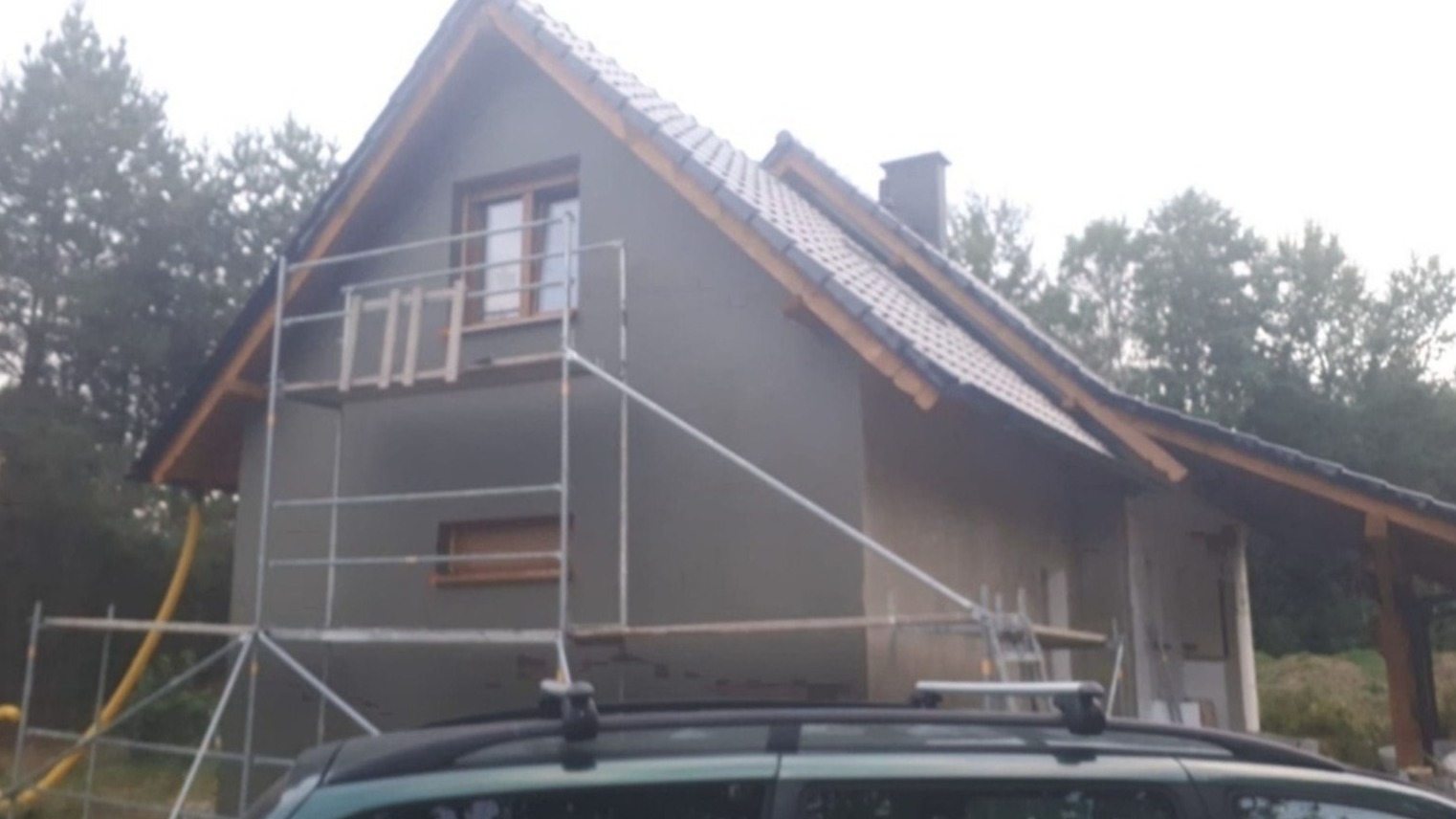 Dom jednorodzinny w trakcie ocieplania elewacji, widoczne rusztowania, szara elewacja i fragment samochodu na pierwszym planie.