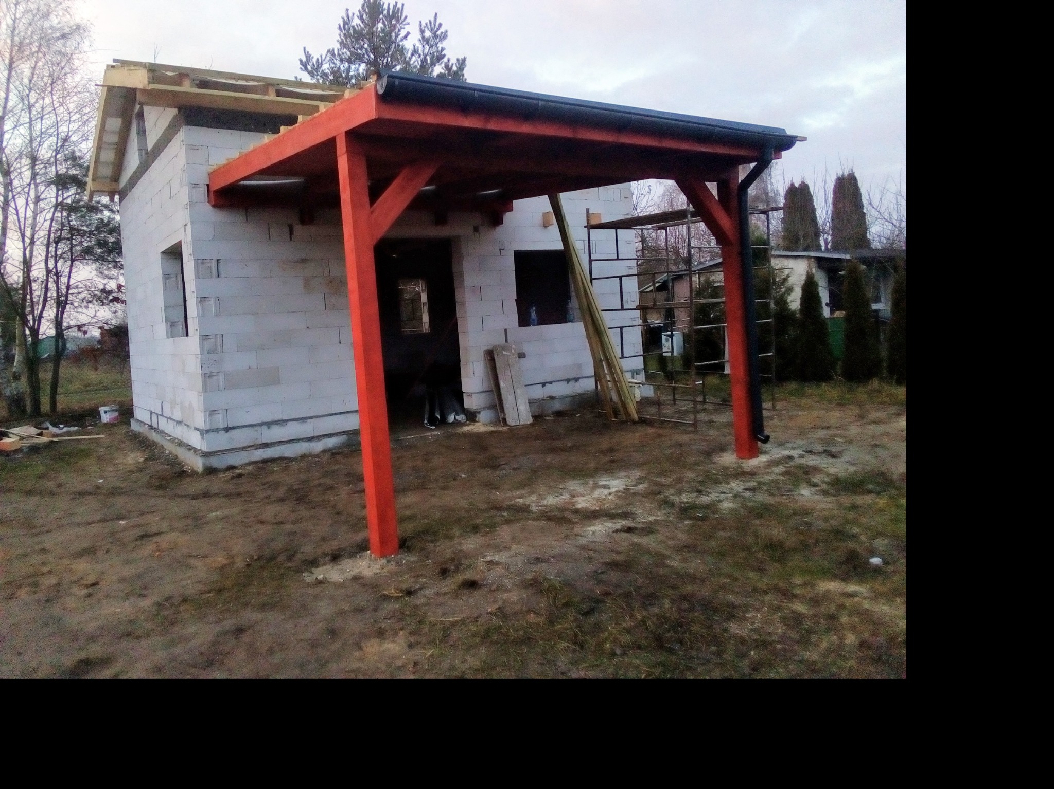 Niedokończona budowa domu z widoczną konstrukcją dachu i nowo zamontowanymi rynnami w kolorze czarnym, na tle pochmurnego nieba i sąsiednich budynków.