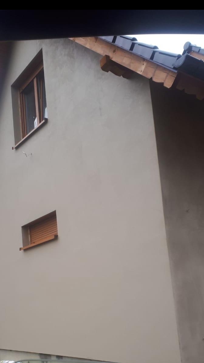 Elewacja domu po malowaniu na kolor beżowy, widoczne dwa okna z drewnianą ramą i roletami, fragment dachu z drewnianymi elementami konstrukcyjnymi.