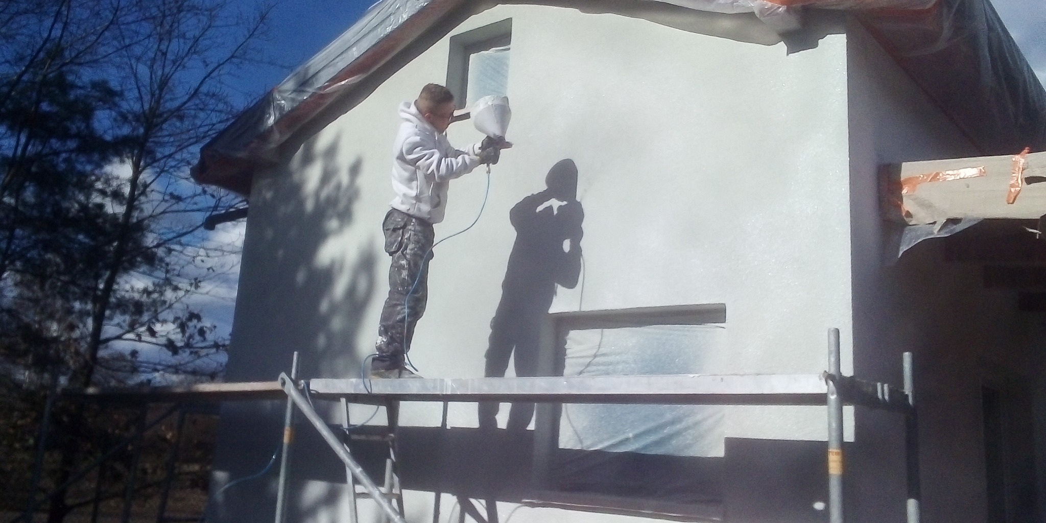 Pracownik w białej bluzie i spodniach moro, stojący na rusztowaniu, aplikuje białą farbę natryskowo na elewację domu. Okna zabezpieczone folią, widoczny cień pracownika na ścianie.