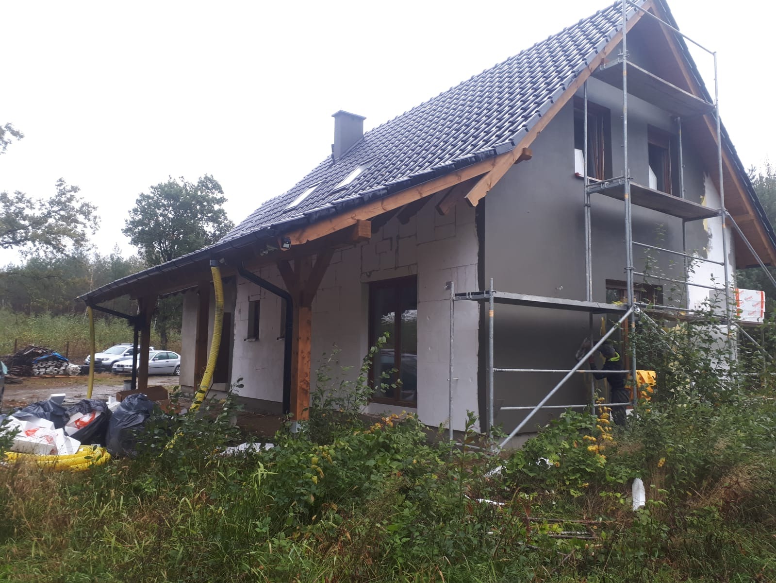 Dom jednorodzinny w trakcie ocieplania; widoczne rusztowania, częściowo otynkowana elewacja, nowy dach z rynnami i żółte rury wentylacyjne.