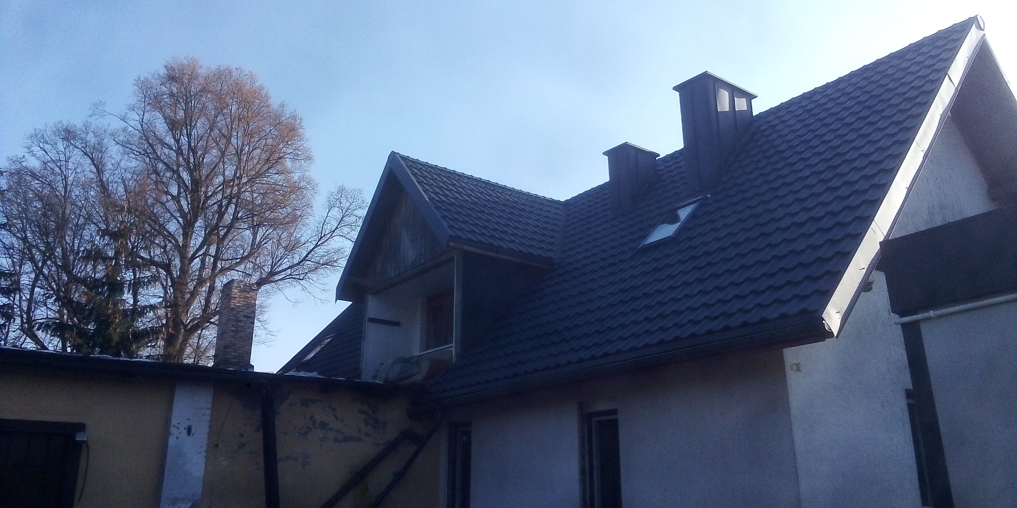 Dom z nowym, ciemnym dachem z blachodachówki, widoczne dwa kominy i okno dachowe, tło stanowi błękitne niebo i drzewa bez liści.
