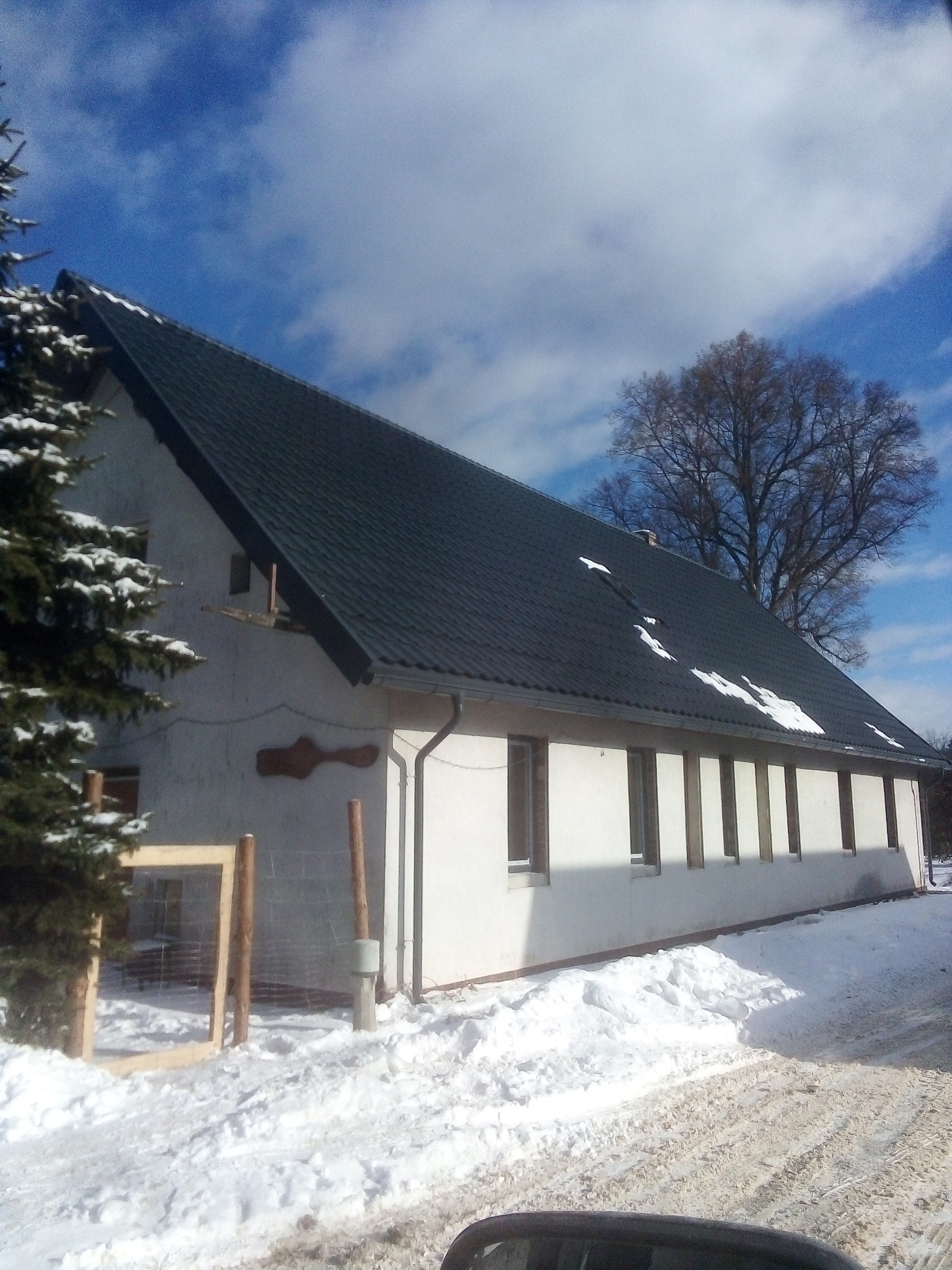 Dwuspadowy dach kryty ciemną dachówką, z fragmentami śniegu, na tle białej ściany budynku z rzędami okien, widok w zimowej scenerii.
