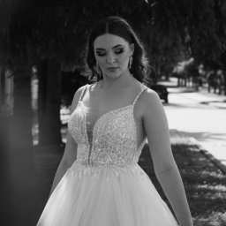 Karol Pałys - Czarno-białe zdjęcie panny młodej w bogato zdobionej sukni z głębokim dekoltem, spoglądającej w dół, z wiszącymi kolczykami, na tle drzew i chodnika.