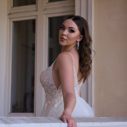 Karol Pałys - Portret młodej kobiety w bogato zdobionej sukni balowej, opierającej się o białą balustradę balkonu, z widocznymi kolczykami i delikatnym makijażem.