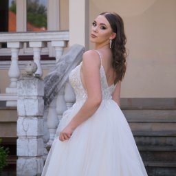 Karol Pałys - Panna młoda w białej sukni z tiulowym dołem, pozująca na tle klasycznej architektury z balustradą, spogląda przez ramię, eksponując długie kolczyki i makijaż.