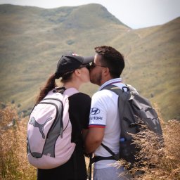 Karol Pałys - Zakochana para turystów w górach, pocałunek na szlaku, plecaki i kije trekkingowe, widok na wzgórza, słoneczny dzień.