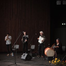 Karol Pałys - Zespół muzyczny na scenie podczas występu: trębacz, dwaj akordeoniści i perkusistka z bębnem, w tle drewniana ściana i reflektory.