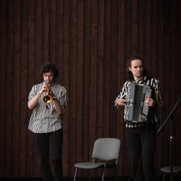 Karol Pałys - Duet muzyczny w pasiastej odzieży: trębacz grający na trąbce i akordeonista grający na akordeonie na scenie z drewnianym tłem.