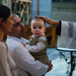 Karol Pałys - Rodzina podczas chrztu dziecka w kościele, ksiądz błogosławi chłopca, tata trzyma syna na rękach, mama stoi obok.