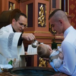 Karol Pałys - Ksiądz w ornacie z haftem roślinnym polewa wodą głowę niemowlęcia trzymanego przez mężczyznę podczas ceremonii w kościele z dekoracyjnym tłem.