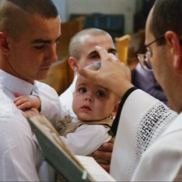 Karol Pałys - Ksiądz polewa wodą głowę dziecka podczas chrztu, ojciec trzyma dziecko w ramionach w kościele.
