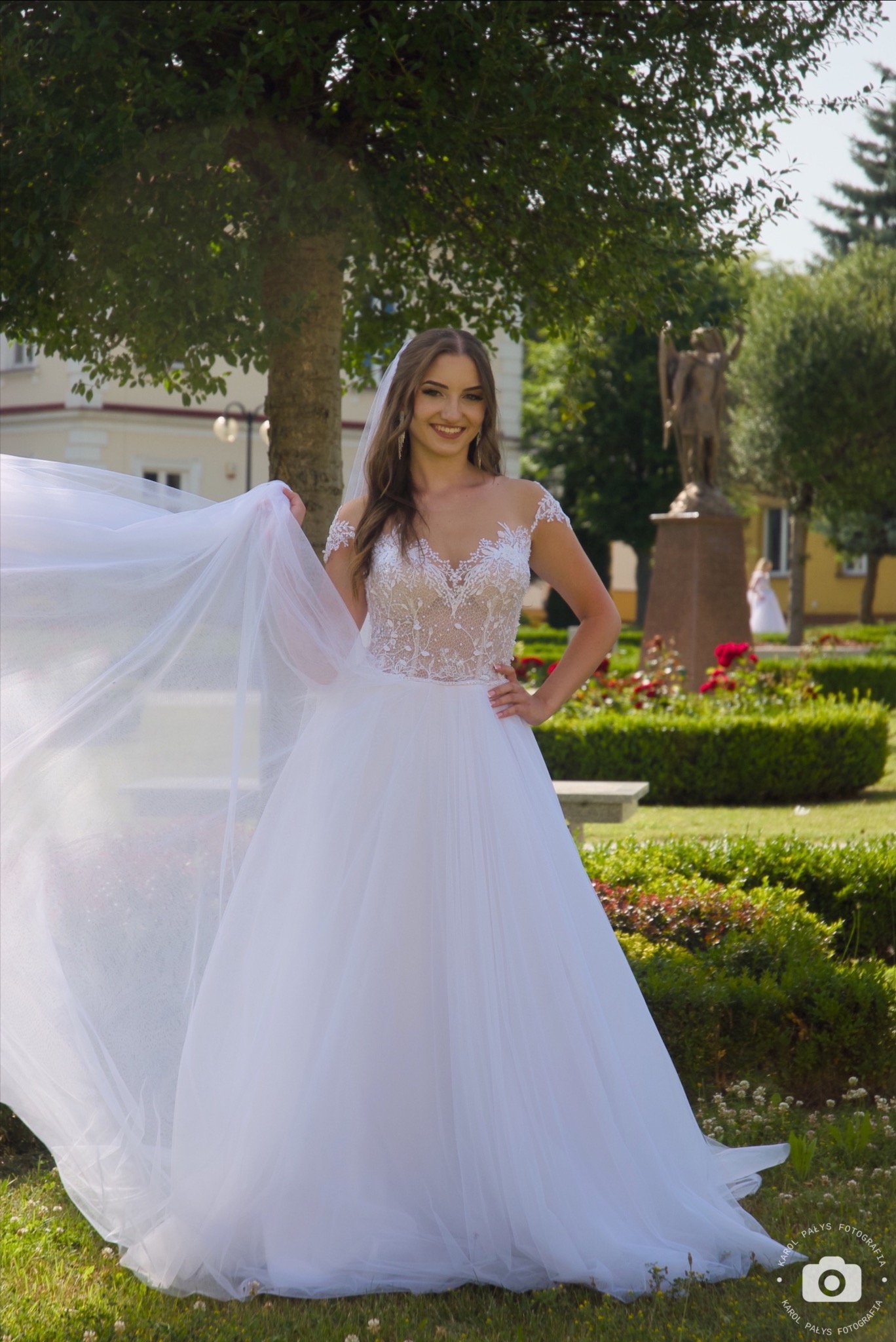 Uśmiechnięta panna młoda w koronkowej sukni, trzymająca welon na tle parku z rzeźbą anioła.