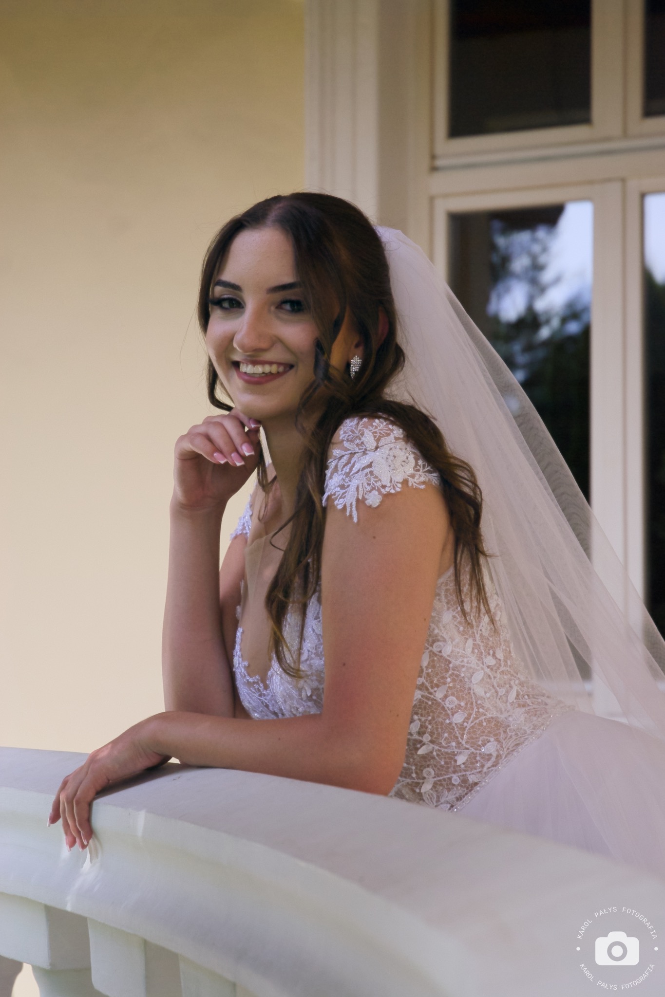Uśmiechnięta panna młoda w koronkowej sukni i welonie, oparta o białą balustradę, z kolczykami z cyrkoniami, na tle okna i jasnej ściany.