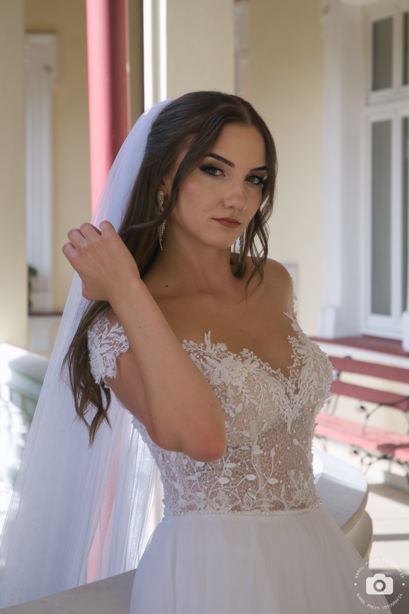 Portret panny młodej w koronkowej sukni z welonem, delikatnie dotykającej włosów, z kolczykami w kształcie kaskady na tle kolumn i okien.