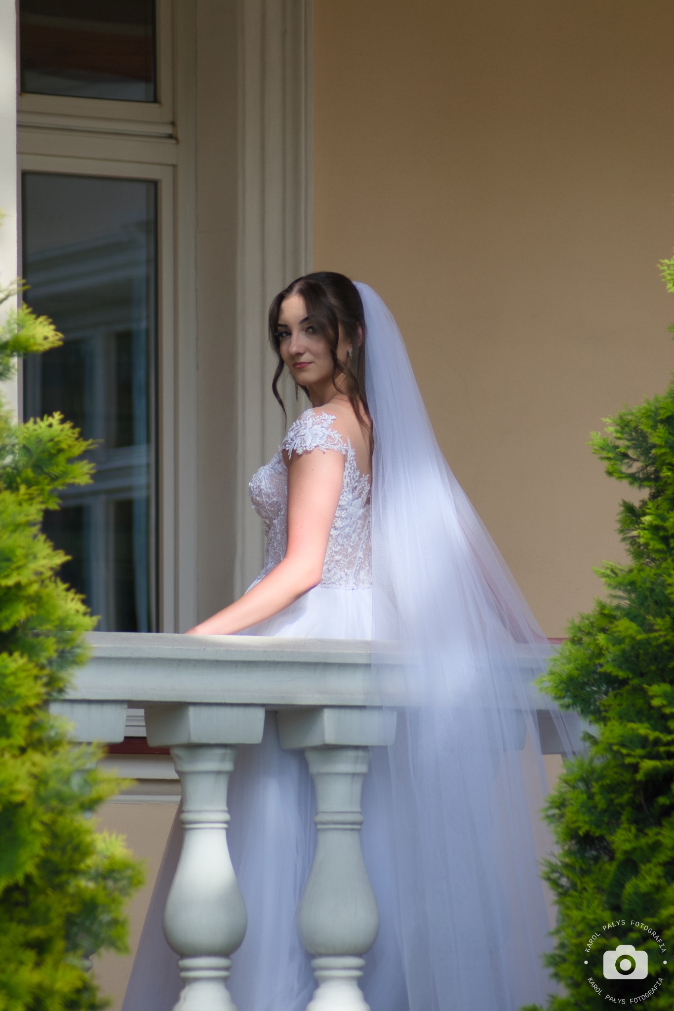 Panna młoda w koronkowej sukni i długim welonie, stojąca na balkonie z białymi balustradami, spogląda przez ramię na fotografa.