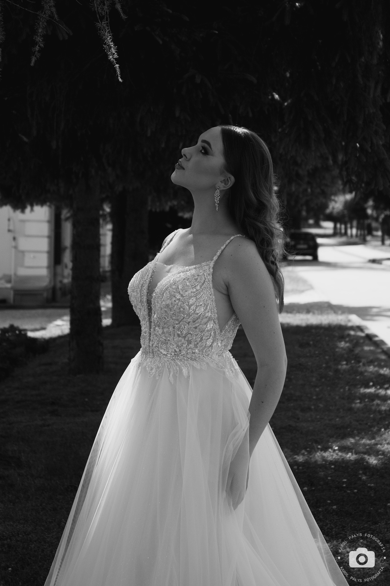 Czarno-białe zdjęcie panny młodej w bogato zdobionej sukni, spoglądającej w górę podczas sesji w Jarosławiu, z widocznym fragmentem drzewa i zabudowań w tle.