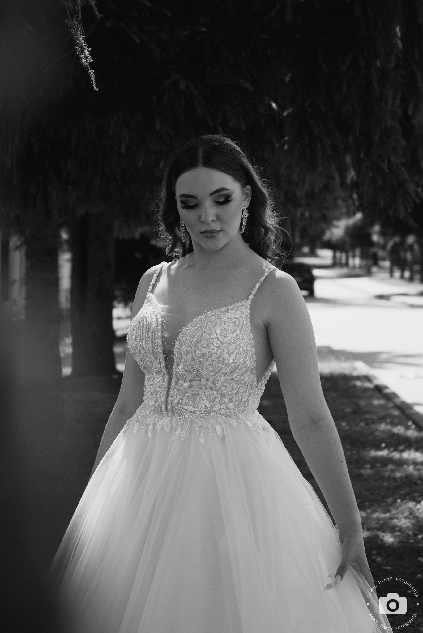 Czarno-białe zdjęcie panny młodej w bogato zdobionej sukni z głębokim dekoltem, spoglądającej w dół, z wiszącymi kolczykami, na tle drzew i chodnika.