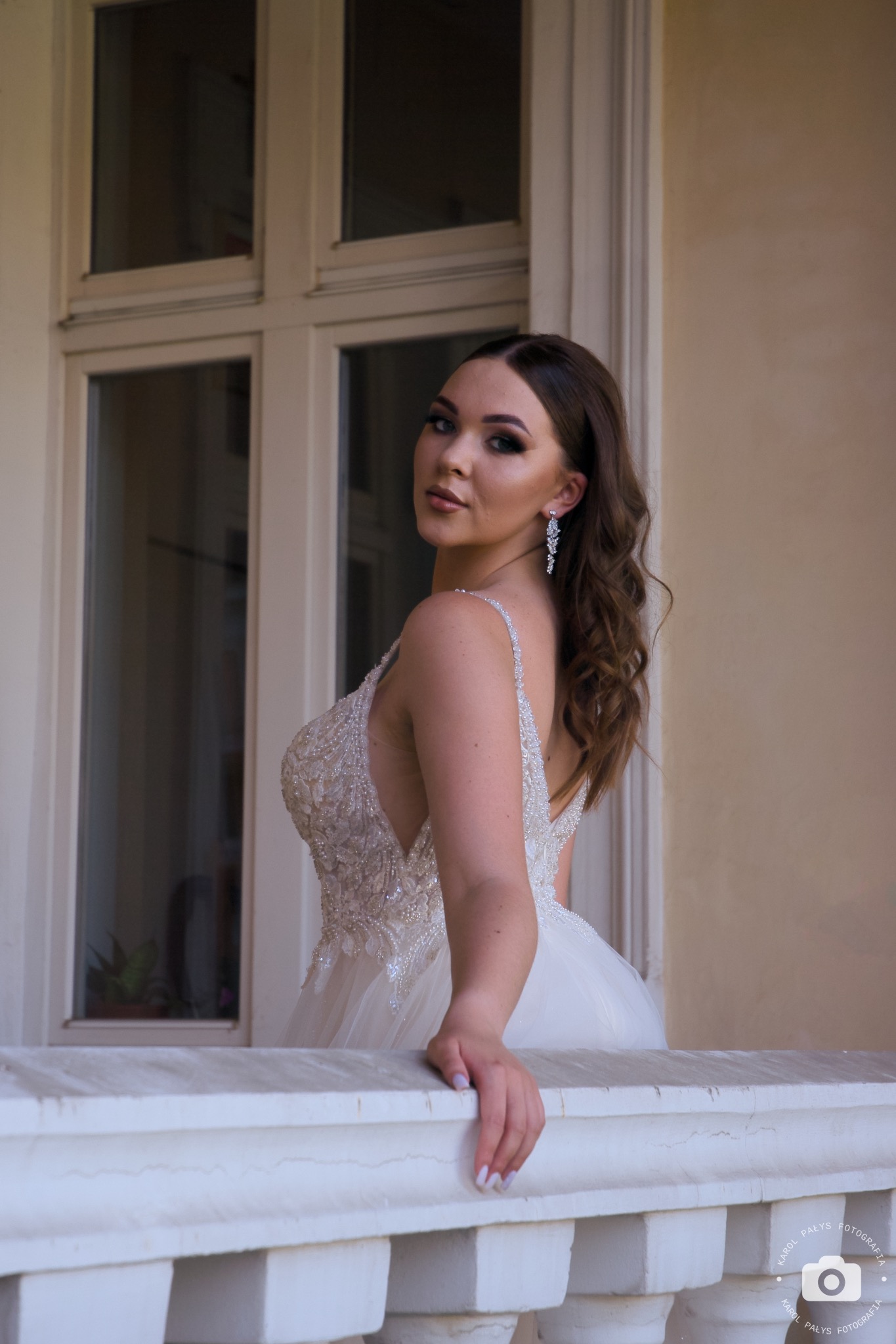 Portret młodej kobiety w bogato zdobionej sukni balowej, opierającej się o białą balustradę balkonu, z widocznymi kolczykami i delikatnym makijażem.