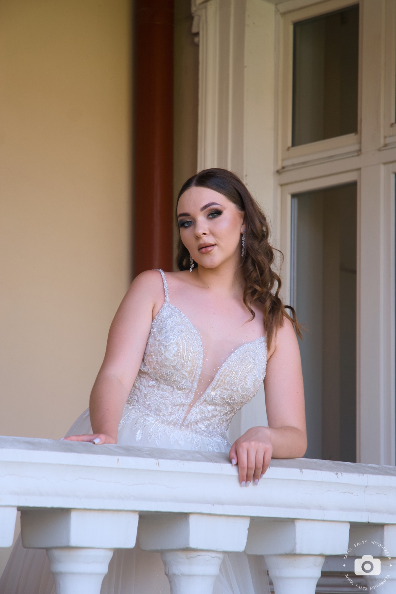 Panna młoda w bogato zdobionej sukni, oparta o białą balustradę balkonu, z delikatnym makijażem i upiętymi włosami, pozuje w naturalnym świetle.