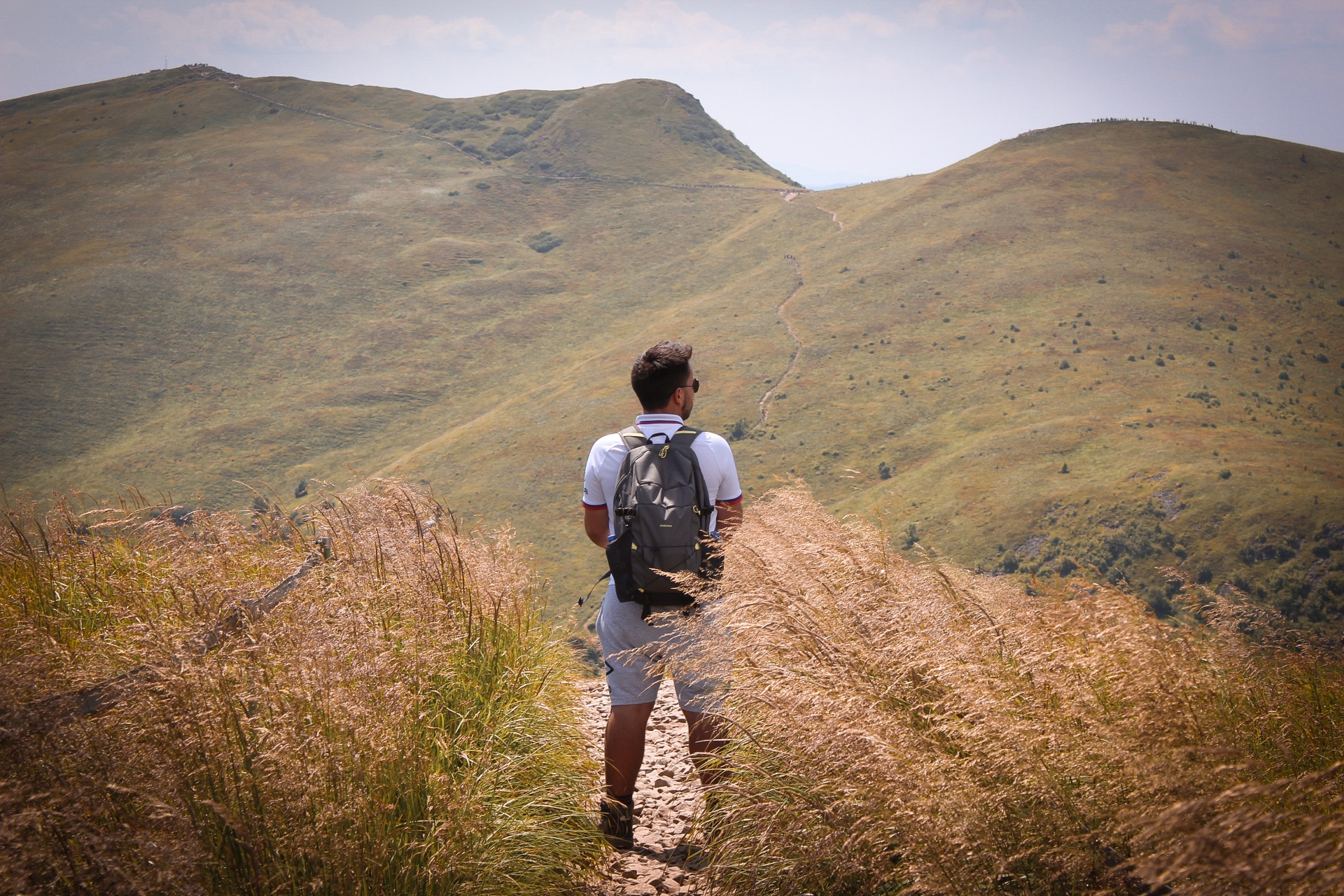 Mężczyzna z plecakiem stoi na szczycie wzgórza porośniętego wysoką trawą, spoglądając na oddalony szlak wiodący na szczyt kolejnego wzniesienia.