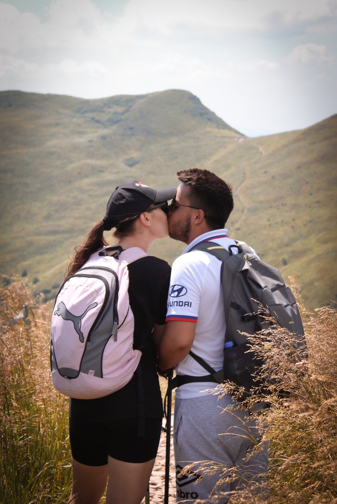 Zakochana para turystów w górach, pocałunek na szlaku, plecaki i kije trekkingowe, widok na wzgórza, słoneczny dzień.