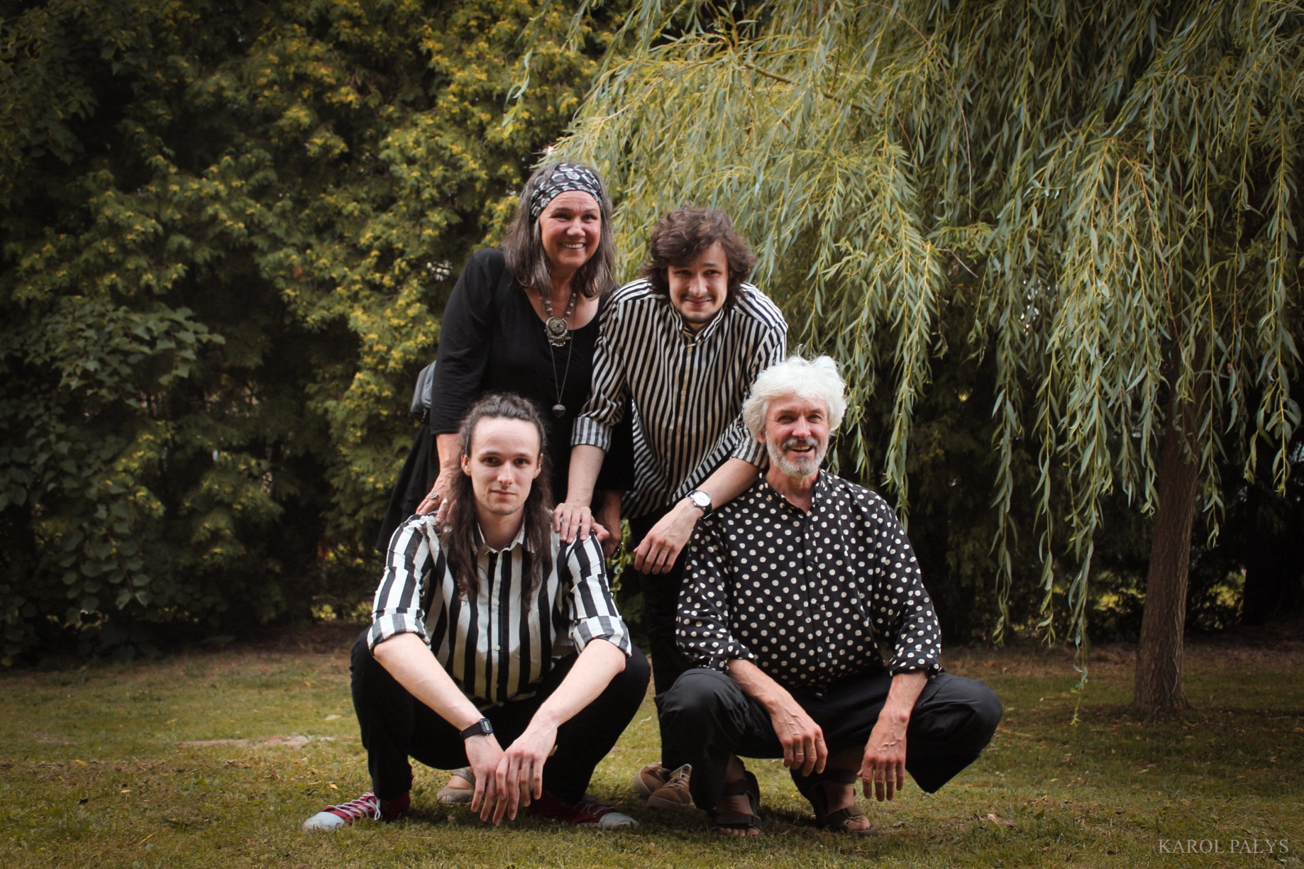 Portret grupy czterech osób w różnym wieku, pozujących na trawniku przed drzewem z wiszącymi gałęziami, w strojach o wzorach w paski i kropki.