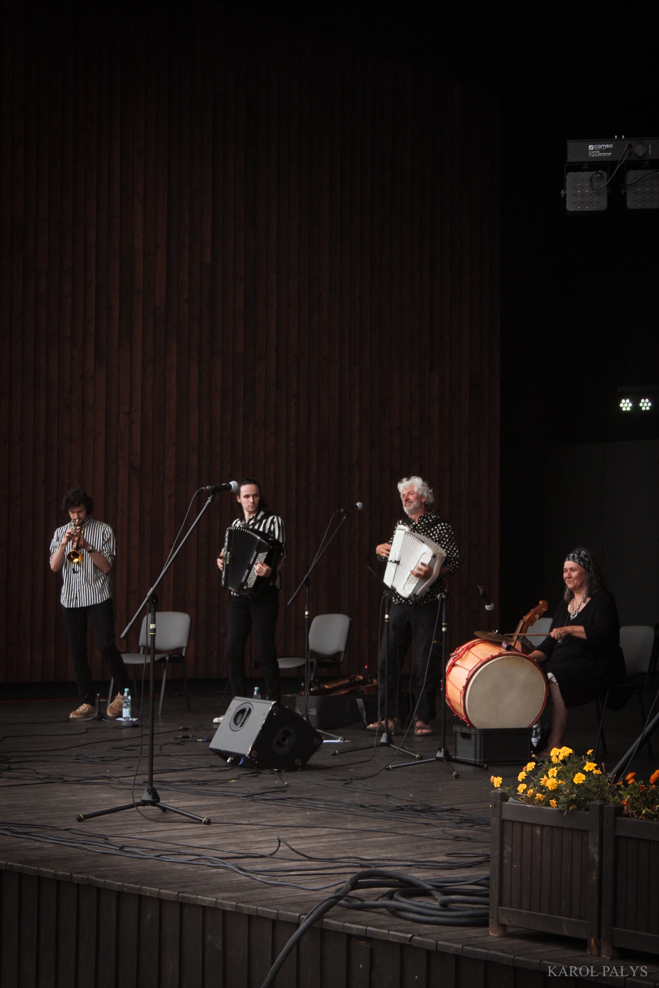 Zespół muzyczny na scenie podczas występu: trębacz, dwaj akordeoniści i perkusistka z bębnem, w tle drewniana ściana i reflektory.