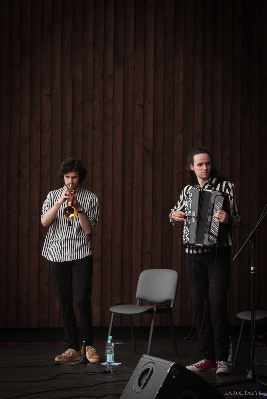 Duet muzyczny w pasiastej odzieży: trębacz grający na trąbce i akordeonista grający na akordeonie na scenie z drewnianym tłem.