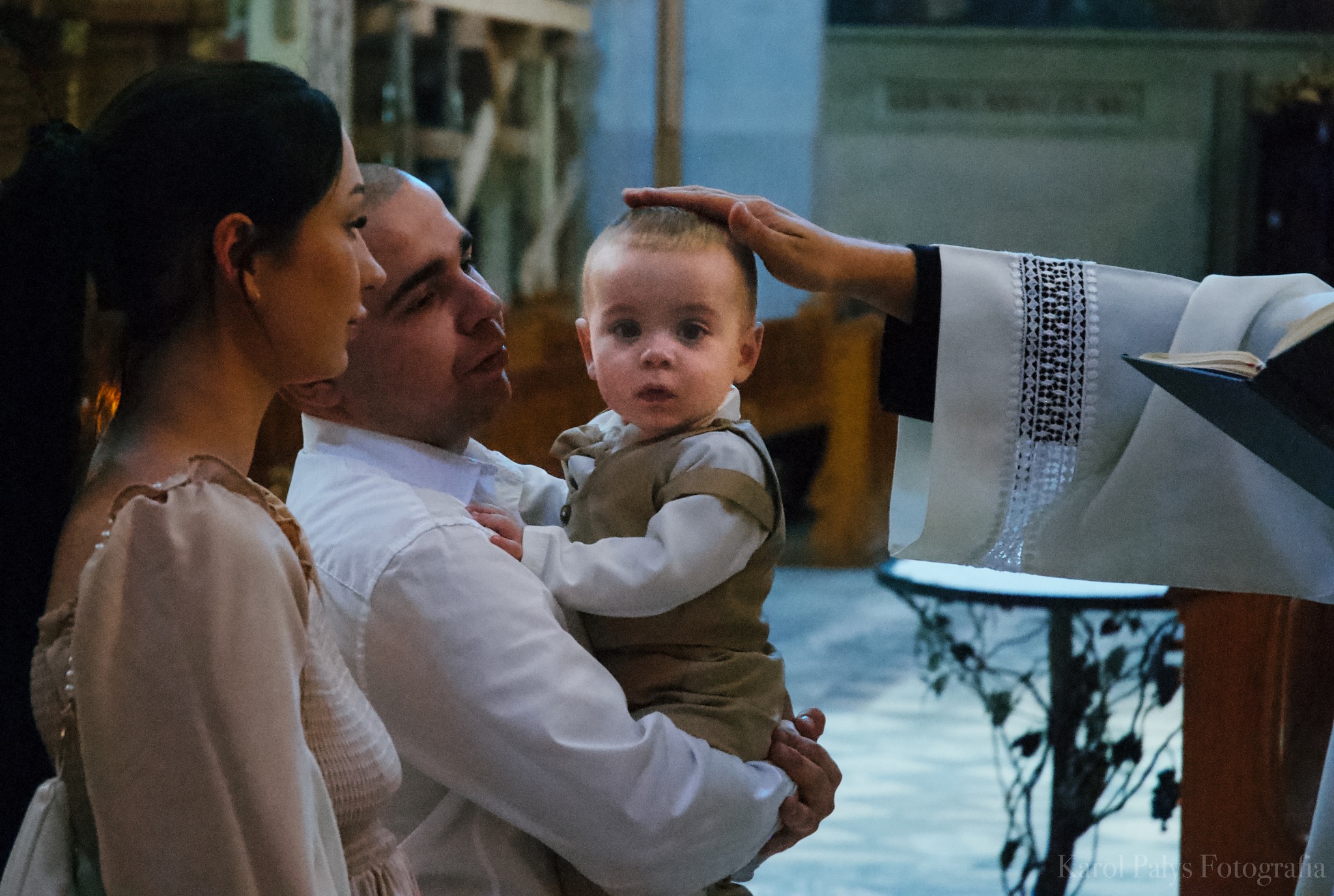 Rodzina podczas chrztu dziecka w kościele, ksiądz błogosławi chłopca, tata trzyma syna na rękach, mama stoi obok.