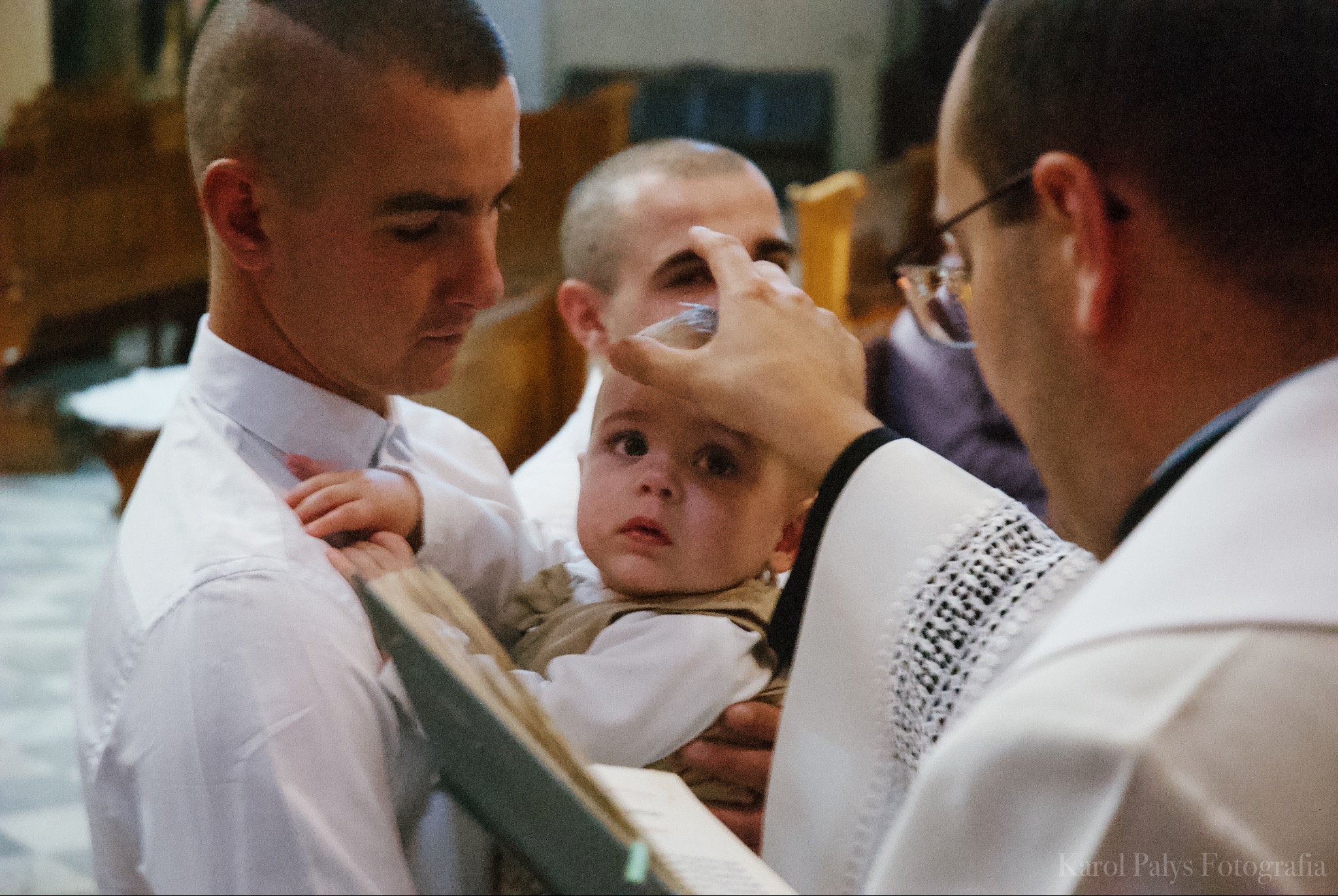 Ksiądz polewa wodą głowę dziecka podczas chrztu, ojciec trzyma dziecko w ramionach w kościele.