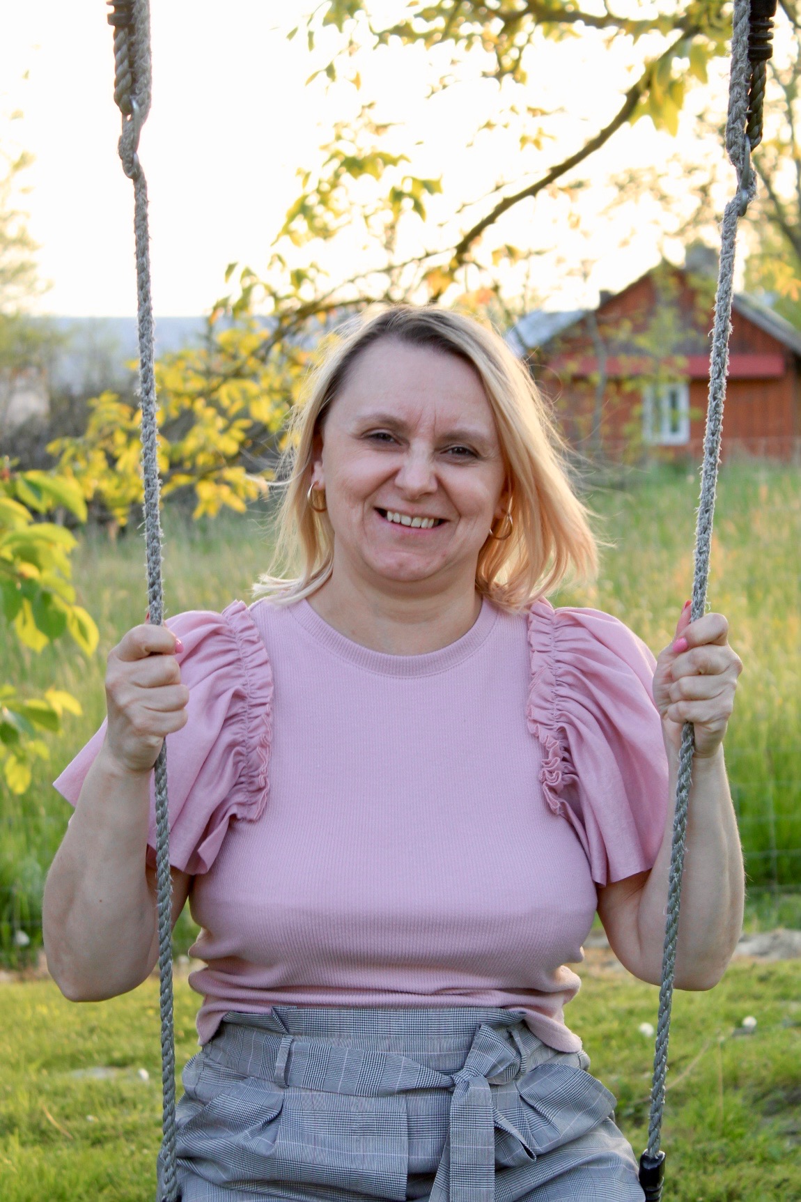 Uśmiechnięta blondynka w różowej bluzce na huśtawce linowej, z domkiem w tle, w ciepłym świetle popołudniowego słońca.