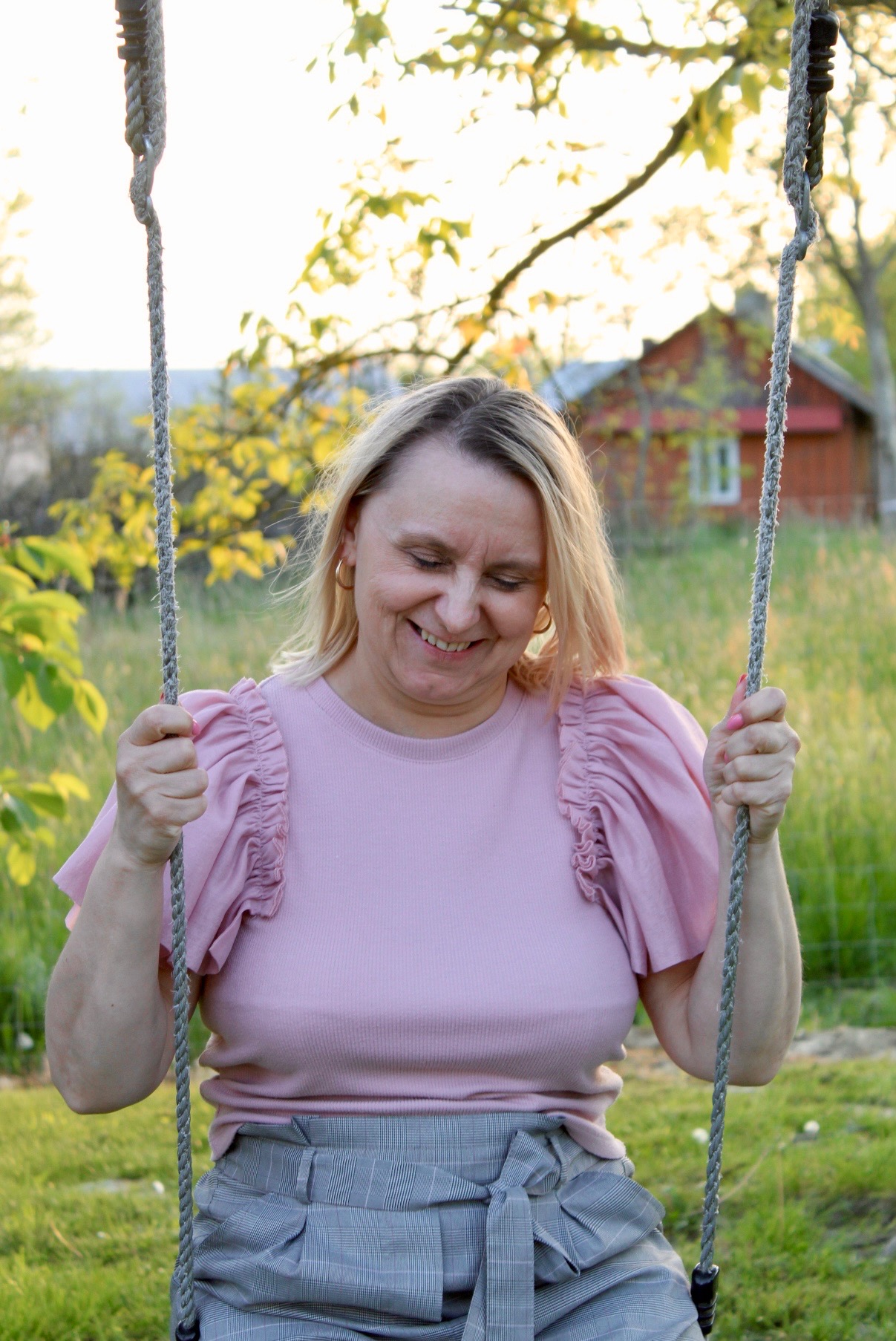 Uśmiechnięta kobieta w różowej bluzce i szarych spodniach na huśtawce linowej w ogrodzie, z domkiem w tle i promieniami słońca przebijającymi przez drzewa.