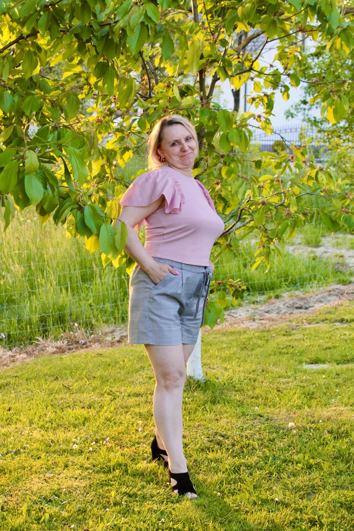 Kobieta w różowej bluzce i szarych spodenkach w kratę pozuje na trawie obok drzewa w słoneczny dzień.
