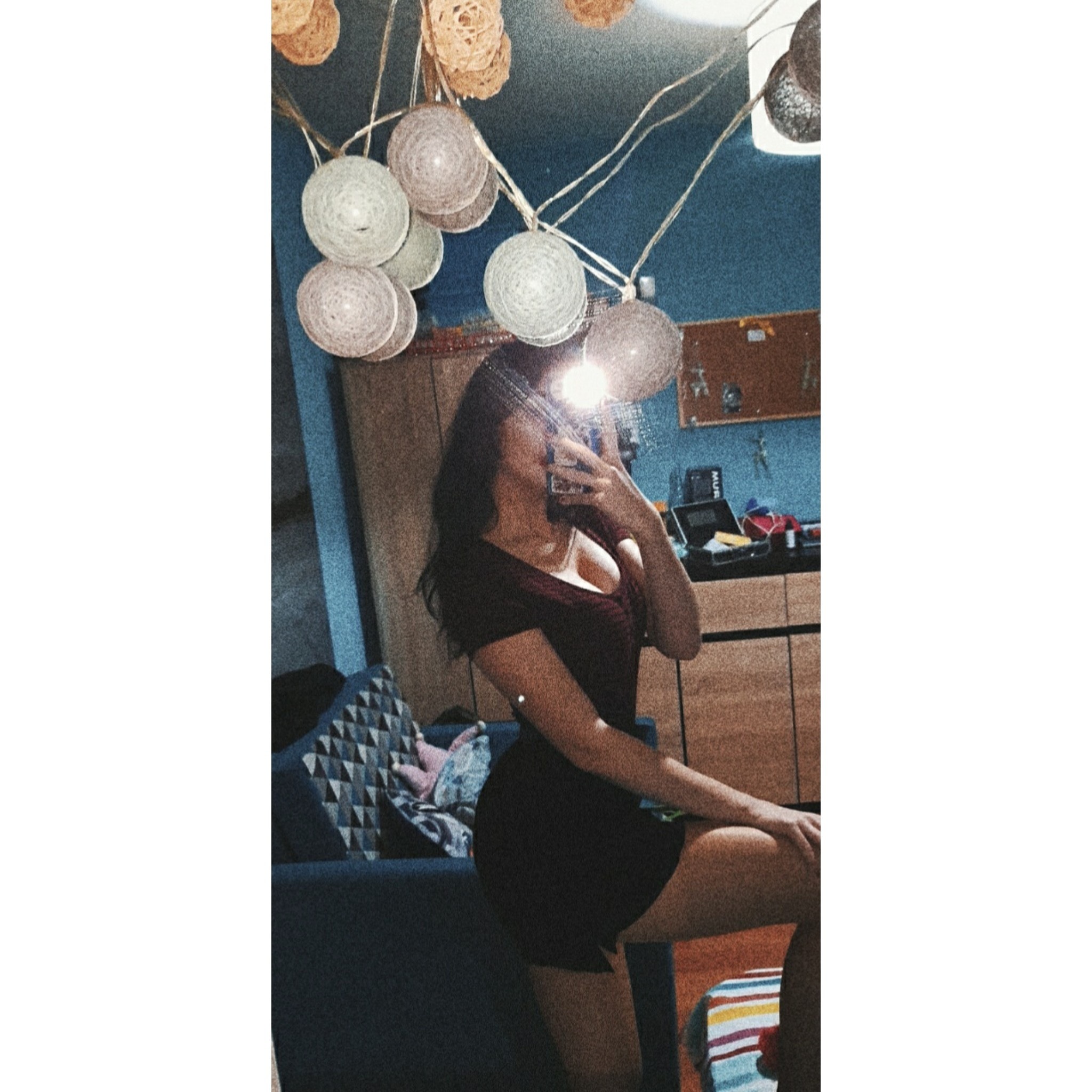 Kobieta w bordowej sukience pozuje przed lustrem, trzymając telefon. W tle widoczne drewniane szafki, niebieska ściana i dekoracyjne lampki cotton balls.