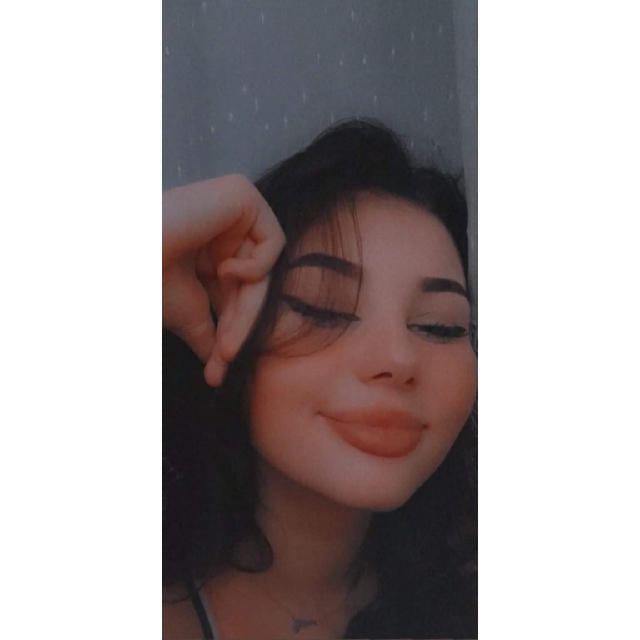 Portret młodej kobiety z ciemnymi włosami i wyrazistym makijażem oczu, opierającej głowę na dłoni, z delikatnym uśmiechem i widocznym naszyjnikiem w kształcie trójkąta.