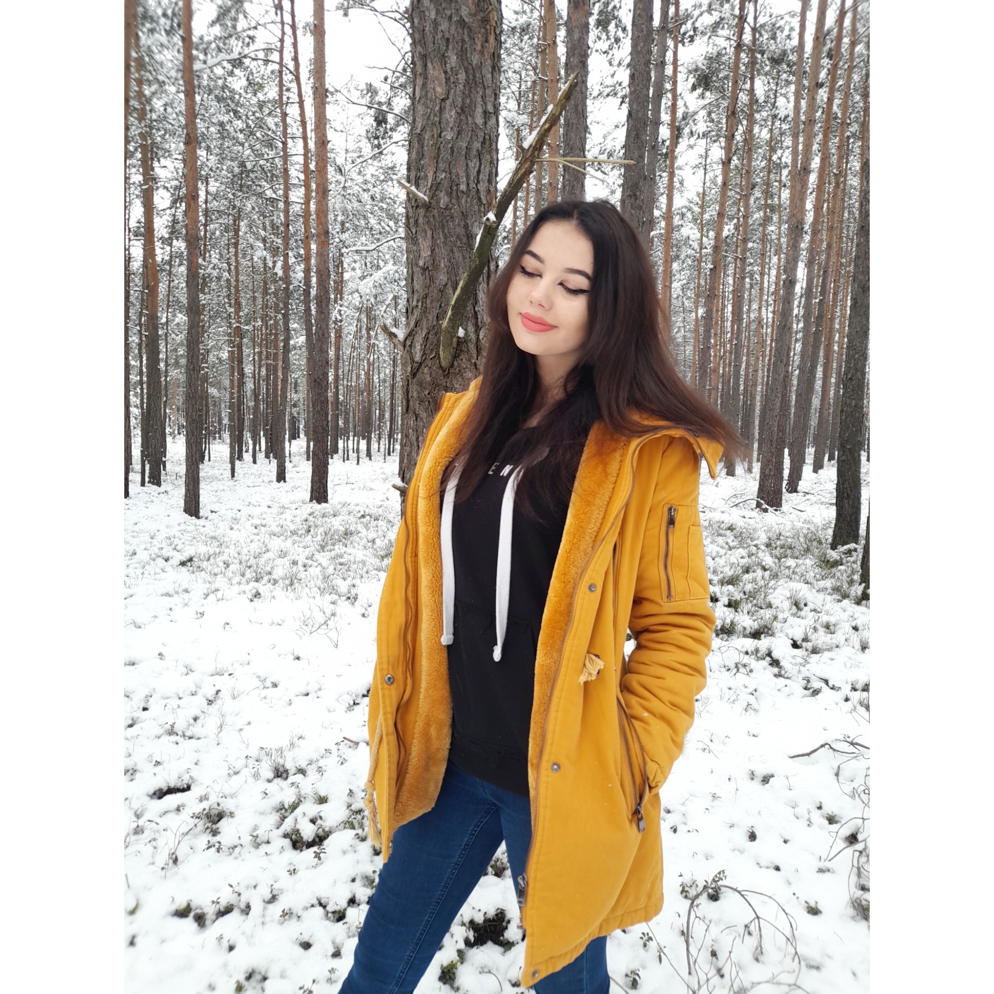 Portret kobiety w żółtej kurtce w zimowym lesie, z przymkniętymi oczami i delikatnym uśmiechem, na tle ośnieżonych drzew.