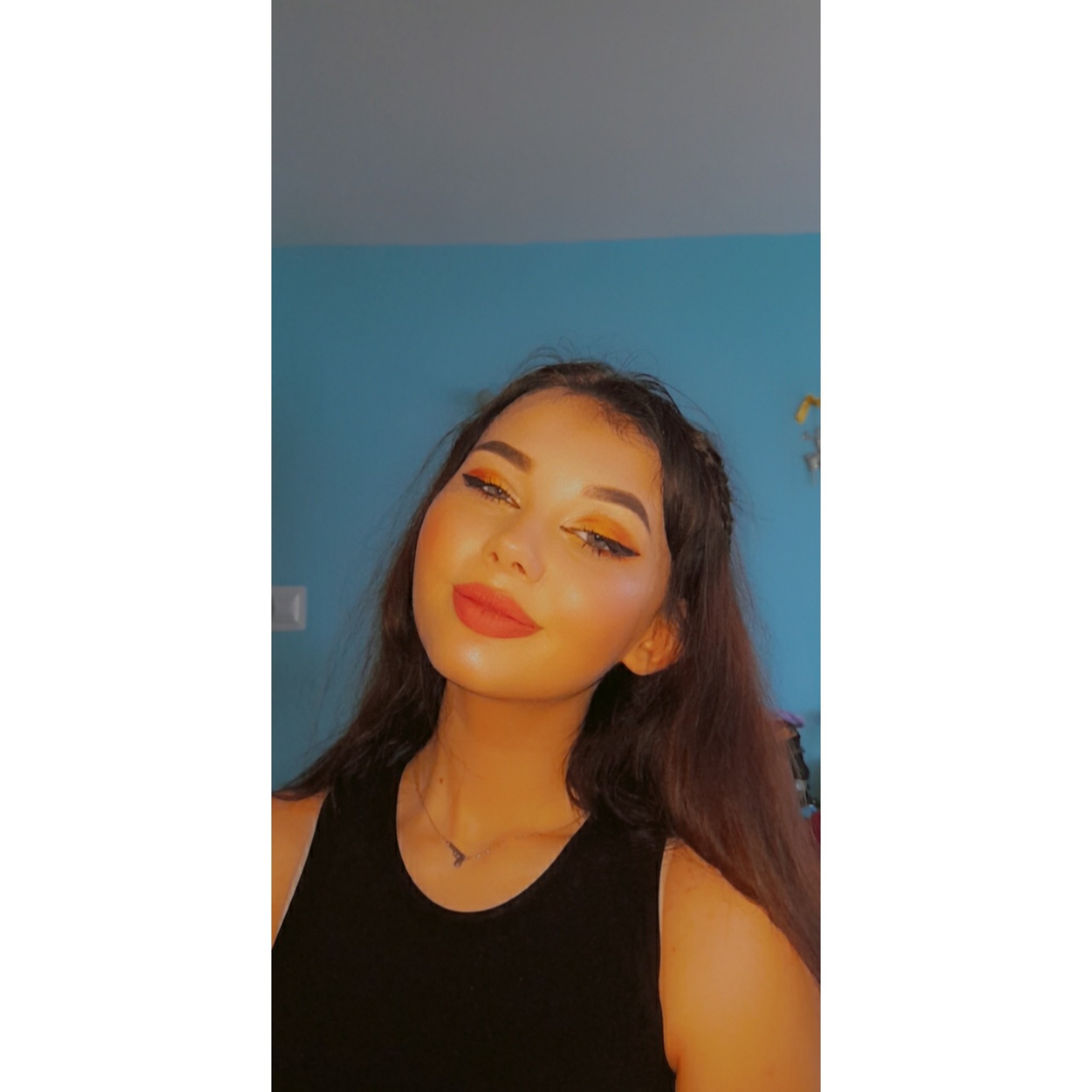 Portret młodej kobiety z mocnym makijażem oczu w odcieniach pomarańczy i czerni, pozującej na tle turkusowej ściany. Widoczny delikatny naszyjnik i czarny top.