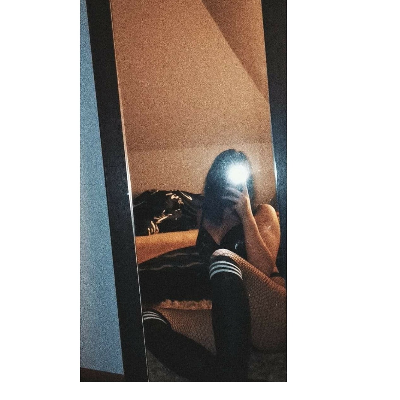 Odbicie w lustrze: Kobieta w bieliźnie i podkolanówkach, robiąca selfie w sypialni z ukośnym sufitem i pościelą w ciemnych kolorach.