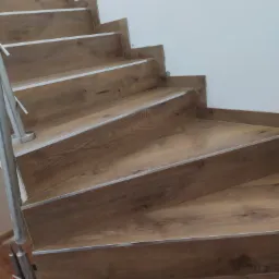 Schody wyłożone panelami drewnopodobnymi z metalowymi listwami zabezpieczającymi krawędzie, widok z dołu, szara poręcz.