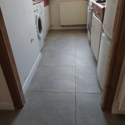 Remonty wykończenia wnętrz - Ułożone szare płytki podłogowe w wąskiej kuchni w Myślenicach, widok z perspektywy wejścia, z widoczną pralką i fragmentem lodówki po bokach.