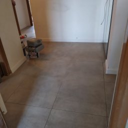 Remonty wykończenia wnętrz - Korytarz z szarymi, kwadratowymi płytkami podłogowymi; widoczny fragment ściany, drzwi i narzędzia pracy w tle.