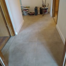Remonty wykończenia wnętrz - Korytarz z nowymi, beżowymi płytkami podłogowymi; w tle widoczne wiadra z farbą, puszki i narzędzia malarskie, sugerujące świeżo zakończone prace.