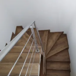 Widok z góry na drewniane schody spiralne z metalową balustradą, widoczne klapki i odkurzacz na dole schodów.