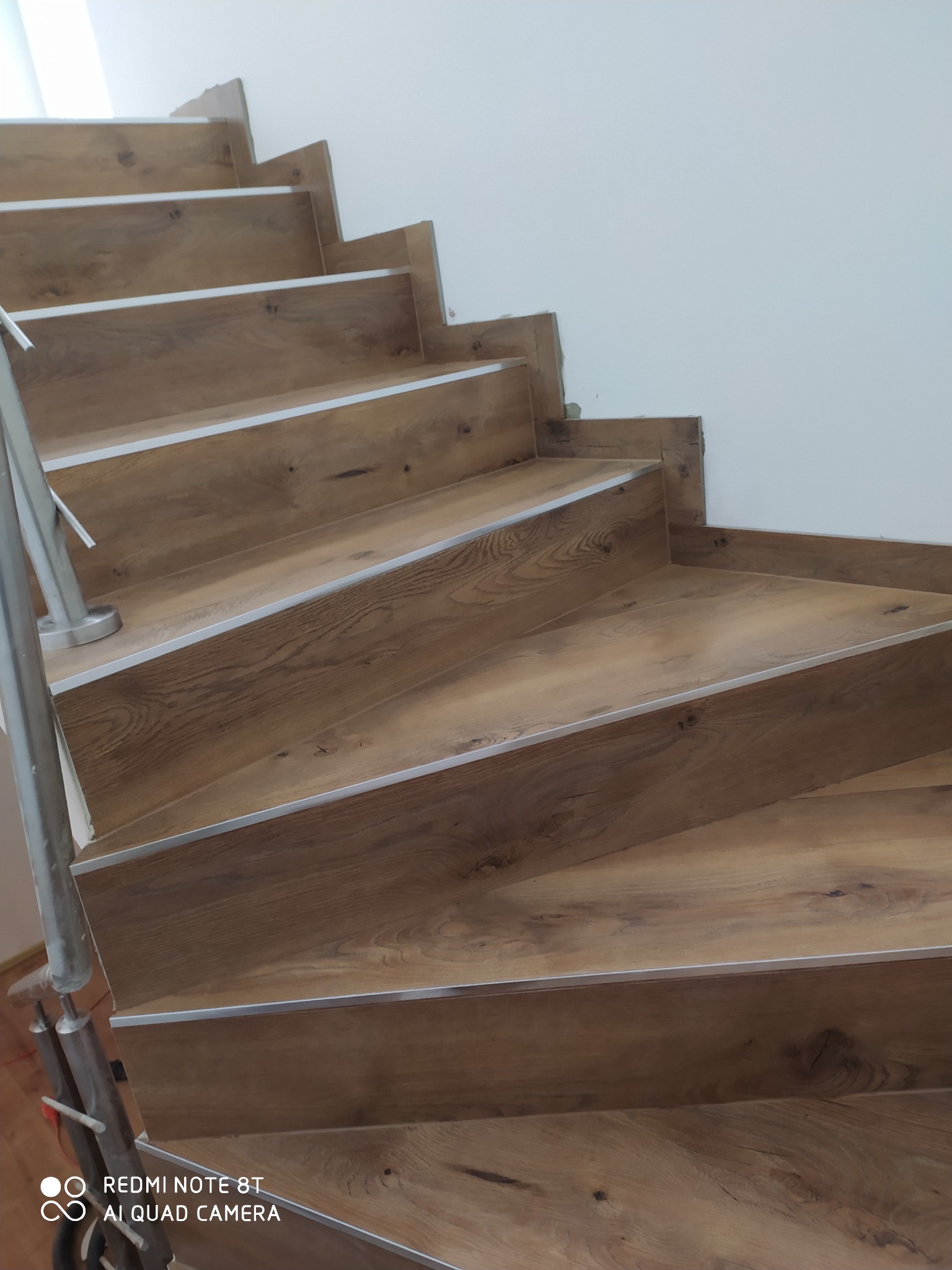 Schody wyłożone panelami drewnopodobnymi z metalowymi listwami zabezpieczającymi krawędzie, widok z dołu, szara poręcz.