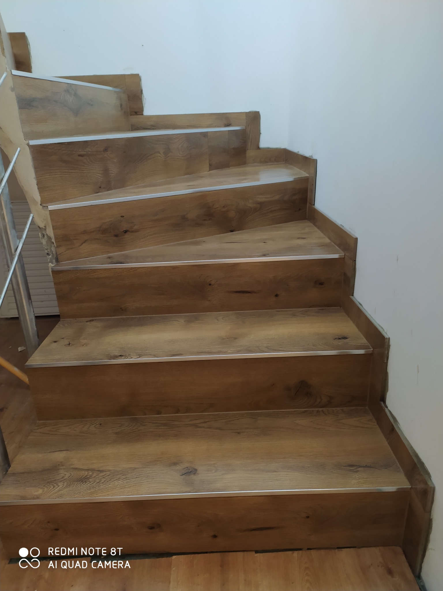 Schody wyłożone panelami imitującymi drewno z metalowymi listwami na krawędziach, widok z dołu, jasne ściany w tle.